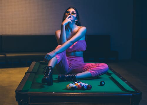 Seductive young female in trendy clothes kissing finger and sitting on billiard table in nightclub