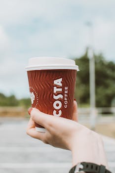 Close-up of a hand holding a Costa Coffee cup in an outdoor setting on a clear day.