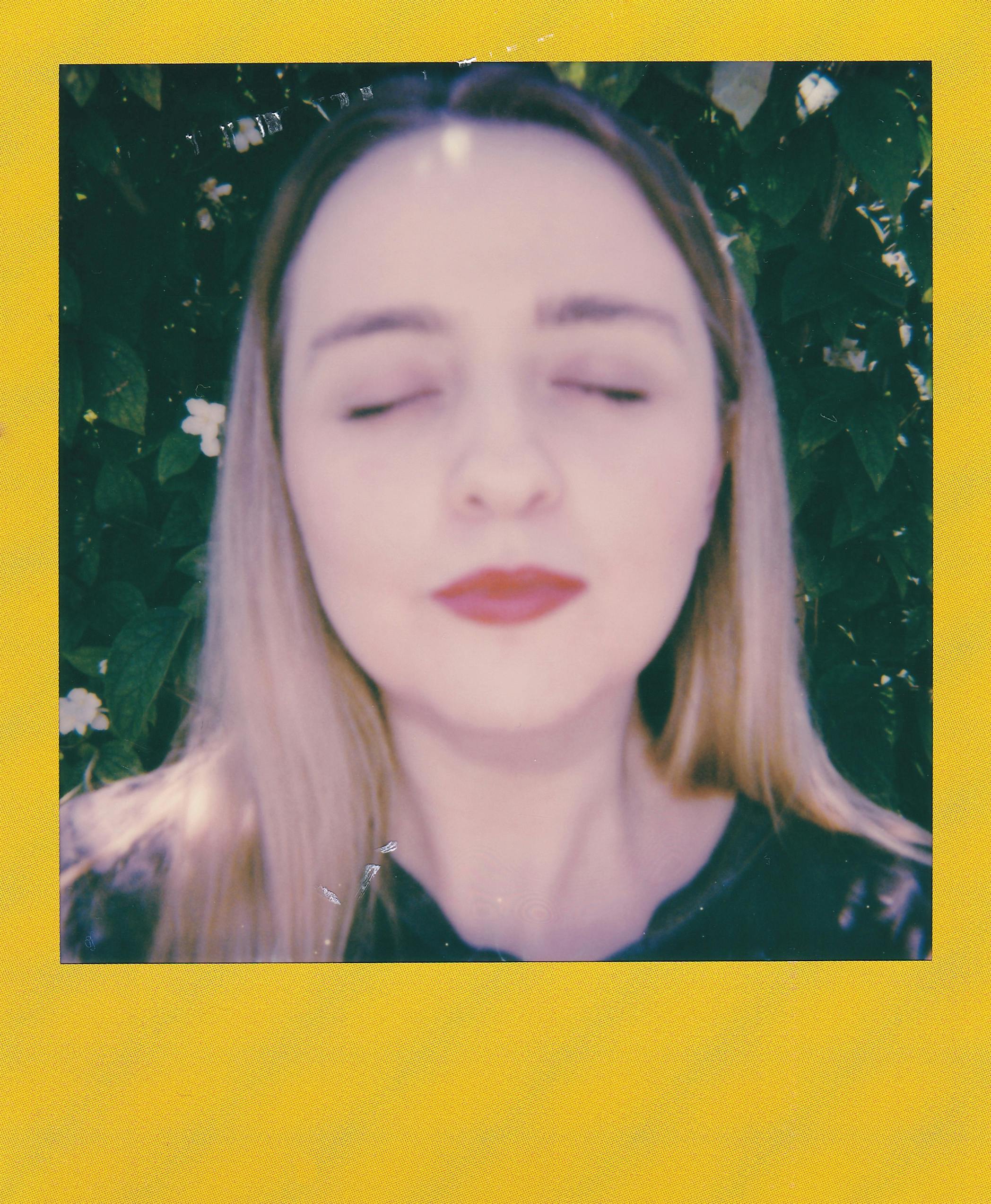 Portrait of a woman with eyes closed, surrounded by green foliage, in a Polaroid frame.