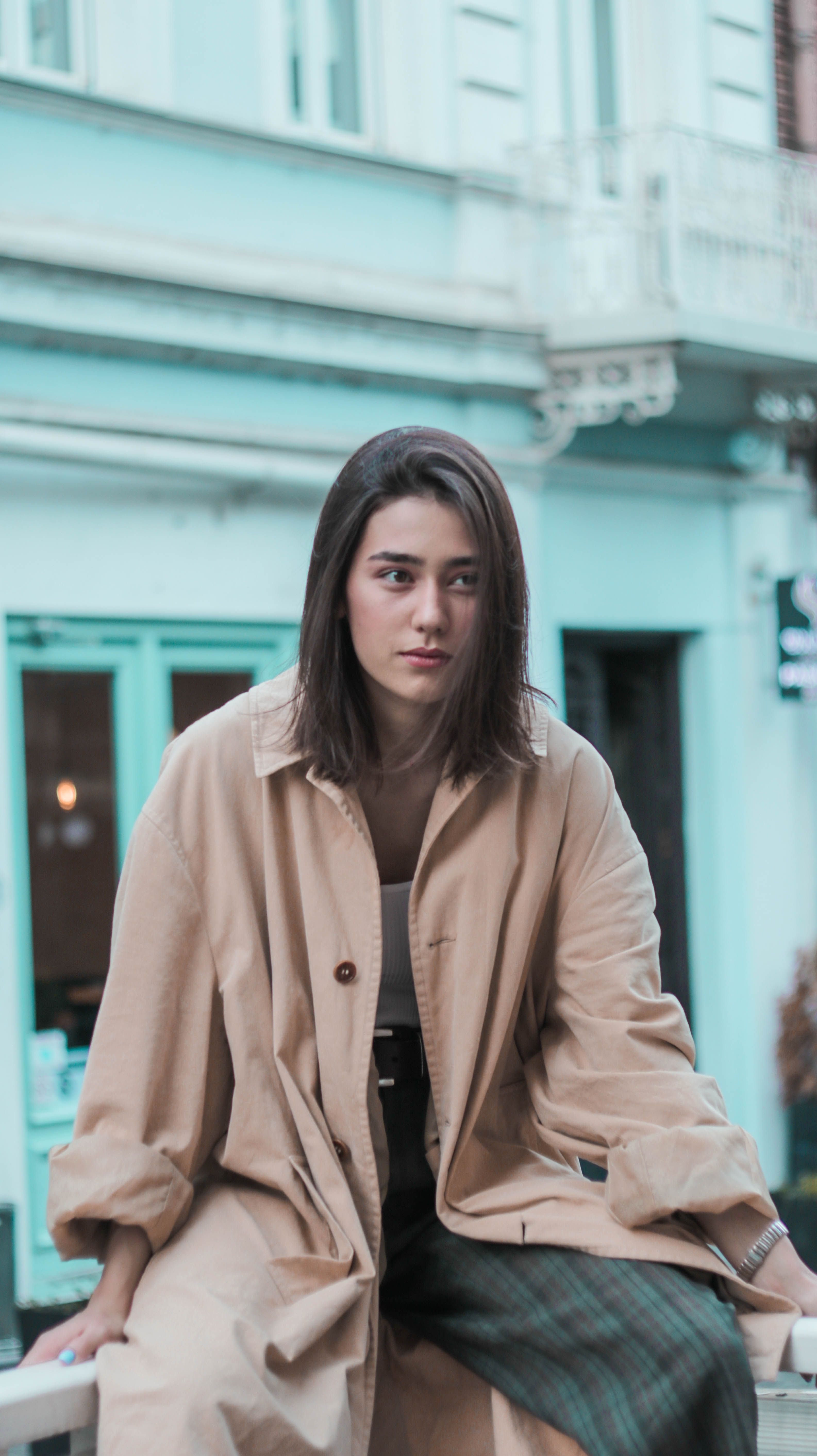 Young calm female in trendy coat sitting on metal railing against blurred building and thoughtfully looking away