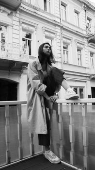 Black and white from below of stylish tired young female in casual outfit sitting on barrier and thoughtfully looking away