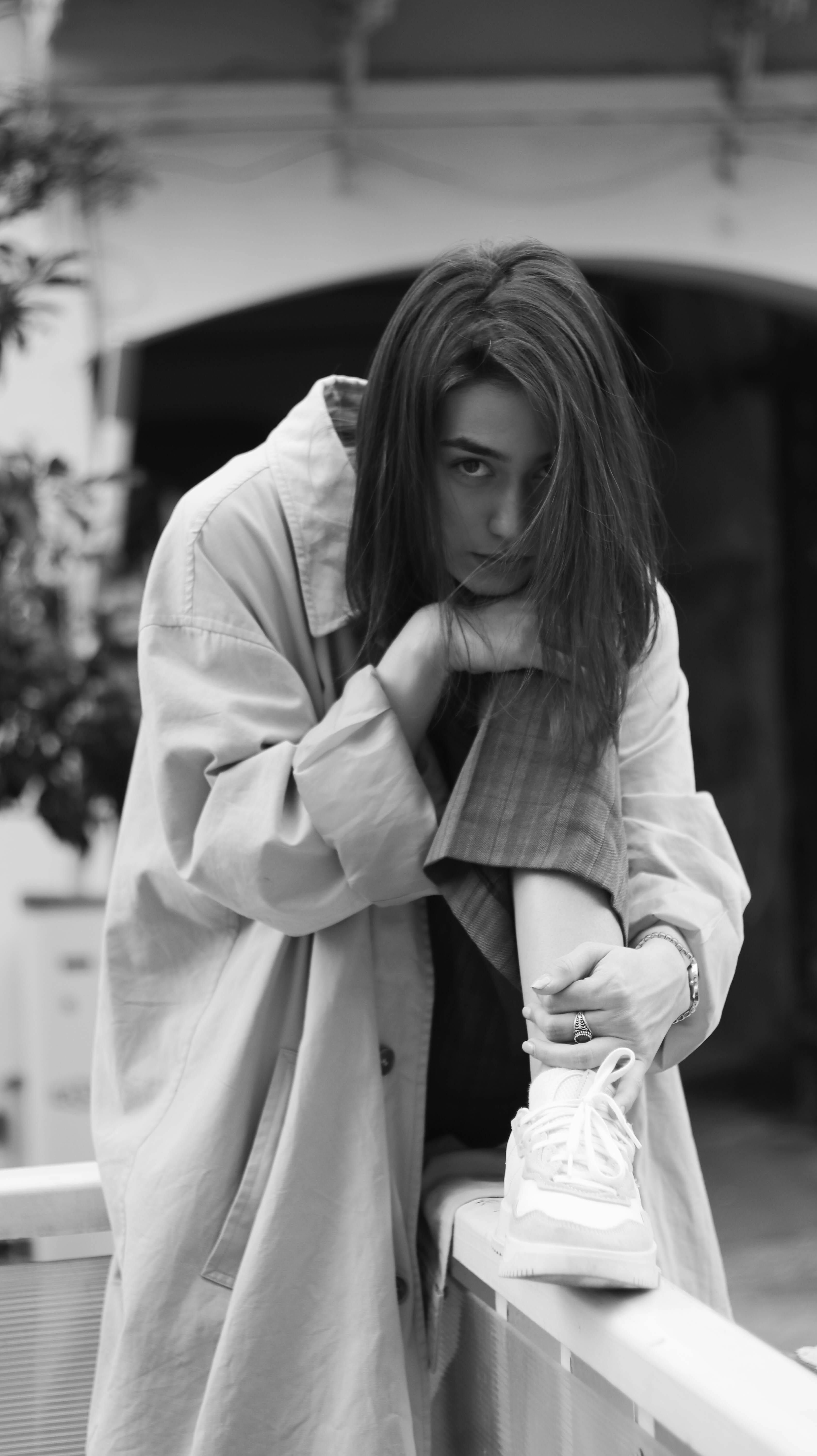 Black and white of calm female in elegant coat and sneakers looking at camera while sitting on border with leg raised and head on knee
