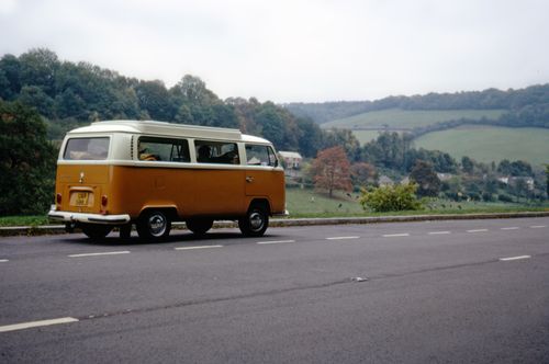 Free Classic orange van parked on a country road beside lush green hills on a cloudy day. Stock Photo