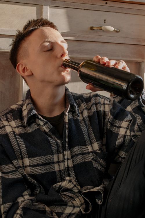 Free A man in a checkered shirt drinks from a bottle indoors, sitting on the floor. Stock Photo