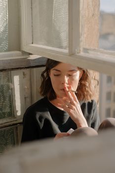 一位妇女在柔和的光线下，以沉思的姿势倚着敞开的窗户吸烟