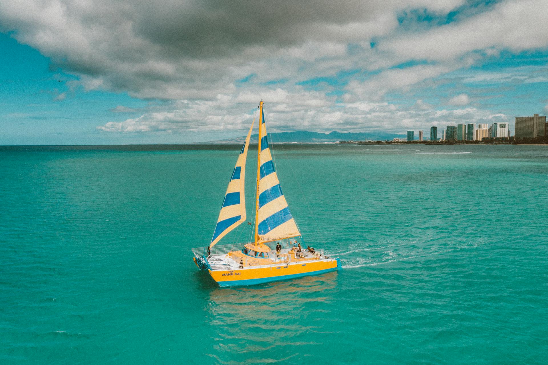 A colorful sailboat cruises on vibrant turquoise waters with city skyline in the distance.