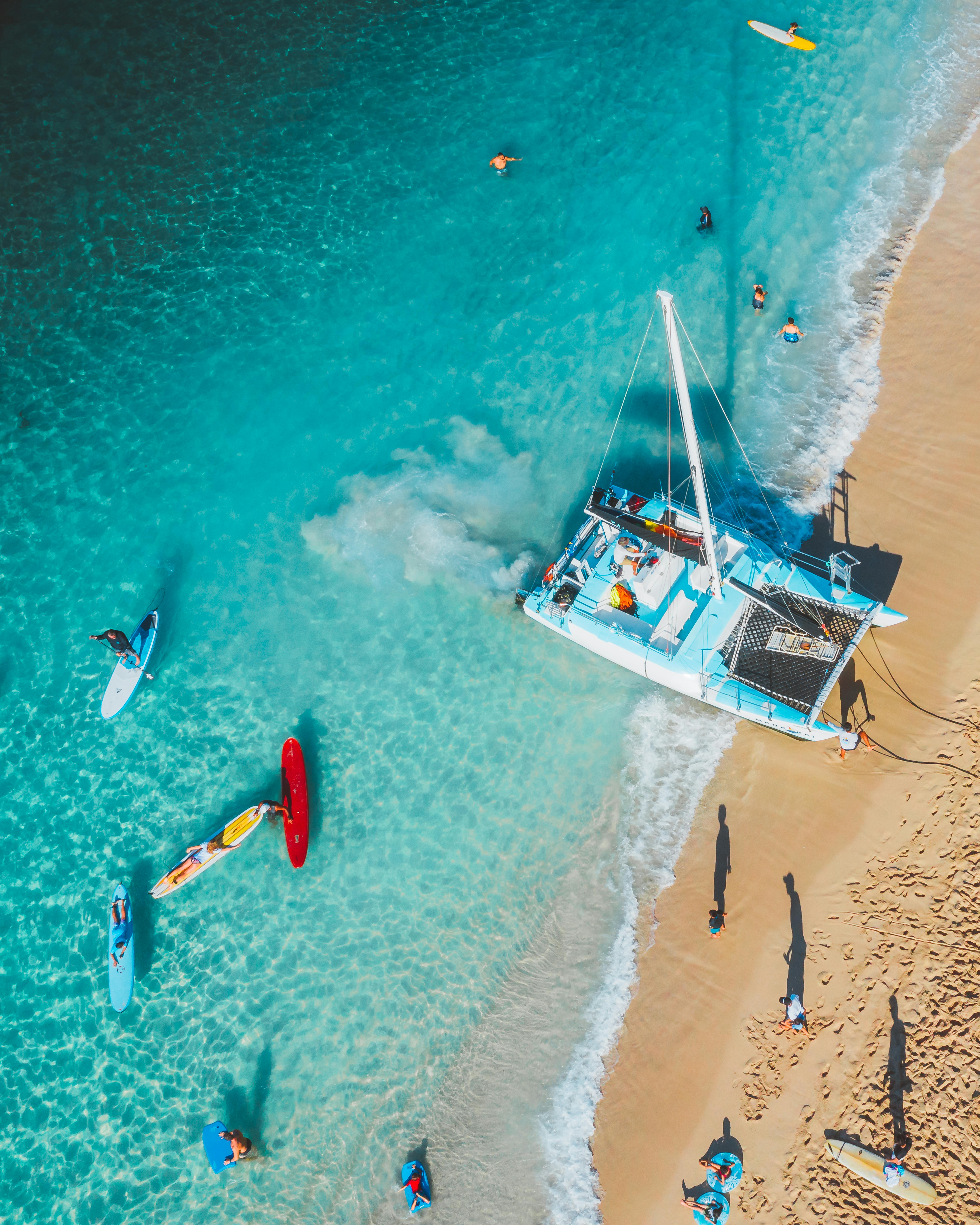 阳光明媚的海滩旁，绿松石水域中，一艘充满活力的双体船正进行空中拍摄，游泳者也在其中