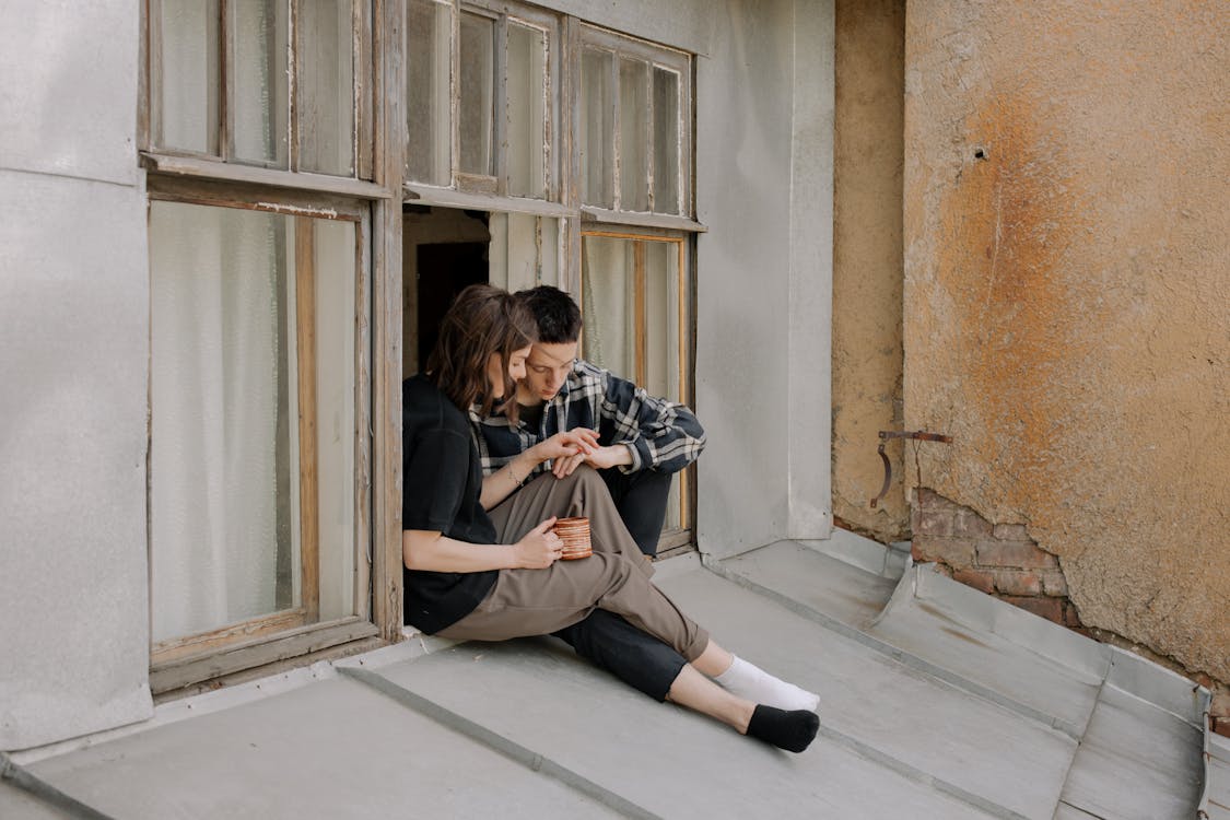 Free A young couple enjoying a peaceful moment on a rooftop window sill, embracing the urban scenery. Stock Photo