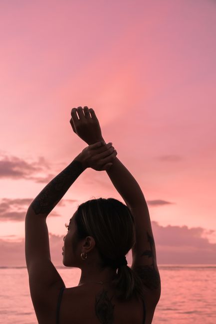 Silhouette of a tattooed woman with arms raised against a pink sunset sky at the beach.