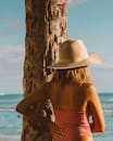 一名戴着帽子和手臂纹身的女子站在阳光明媚的海滩上的一棵棕榈树旁