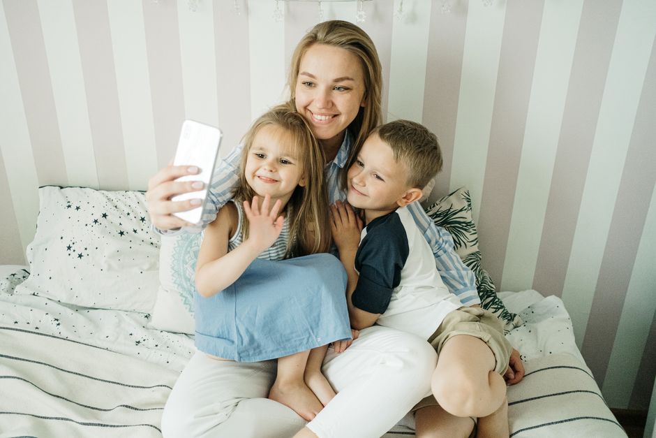 A cheerful mother takes a selfie with her two children on the bed, capturing a bonding moment.