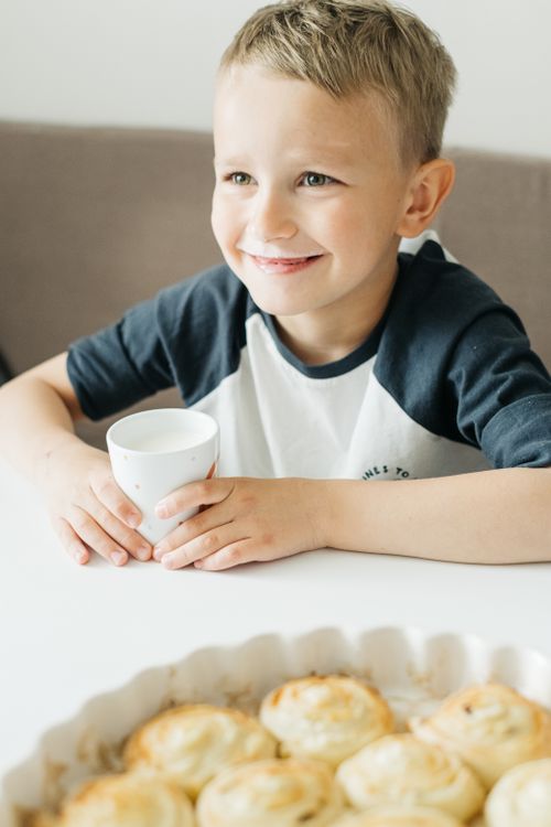 Free Young boy smiling with a cup and cinnamon rolls on the table, enjoying breakfast at home. Stock Photo