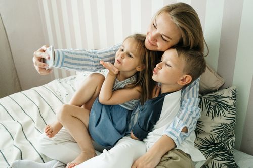 Free A mother with her son and daughter taking a playful selfie on a bed, showcasing family bonding. Stock Photo