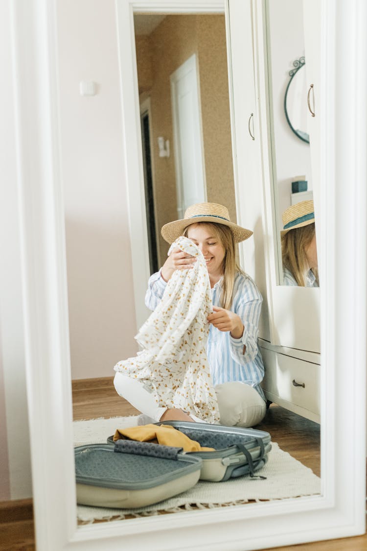 A Woman Wearing A Sun Hat Packing Clothes In A Suitcase