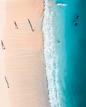 Drone shot capturing people enjoying sunny beach with turquoise waters and sandy shore.