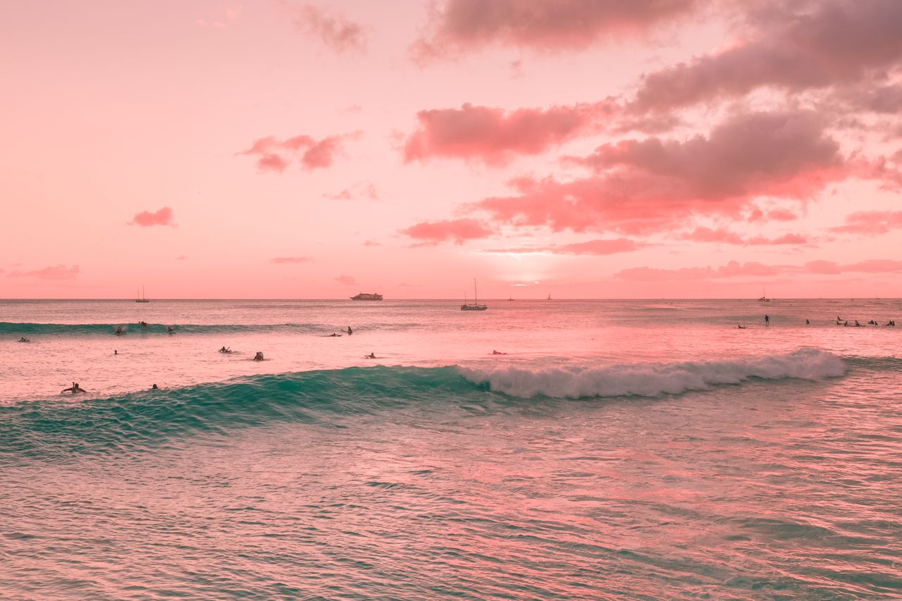 surfers ride gentle ocean waves under a dramatic pink sunset sky surro
