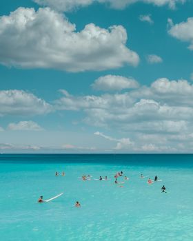 A vibrant scene of surfers and swimmers enjoying a sunny day at a tranquil beach with clear waters and a blue sky.