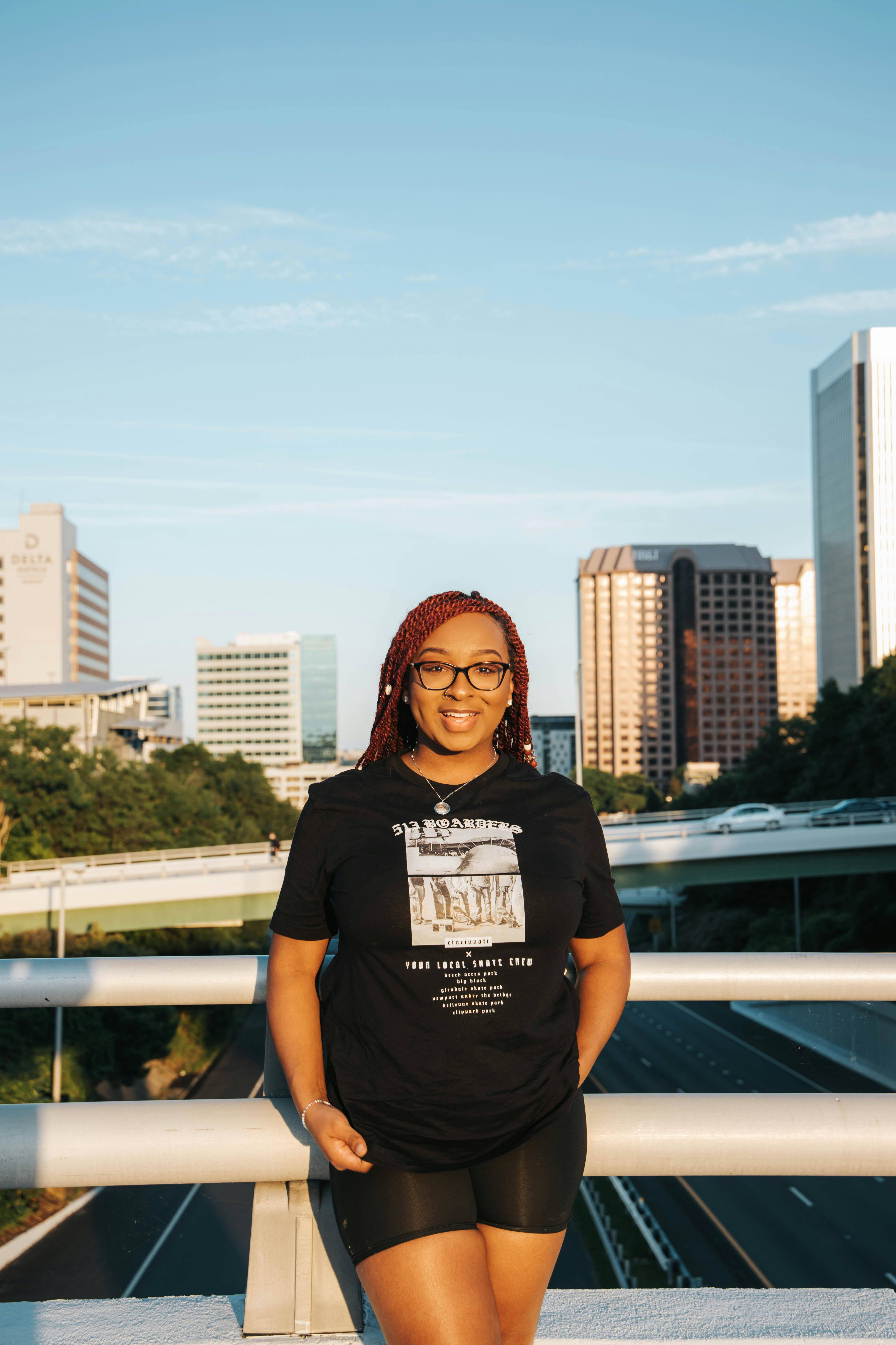 A cheerful woman in a black t-shirt and glasses poses with an urban skyline backdrop during the day.