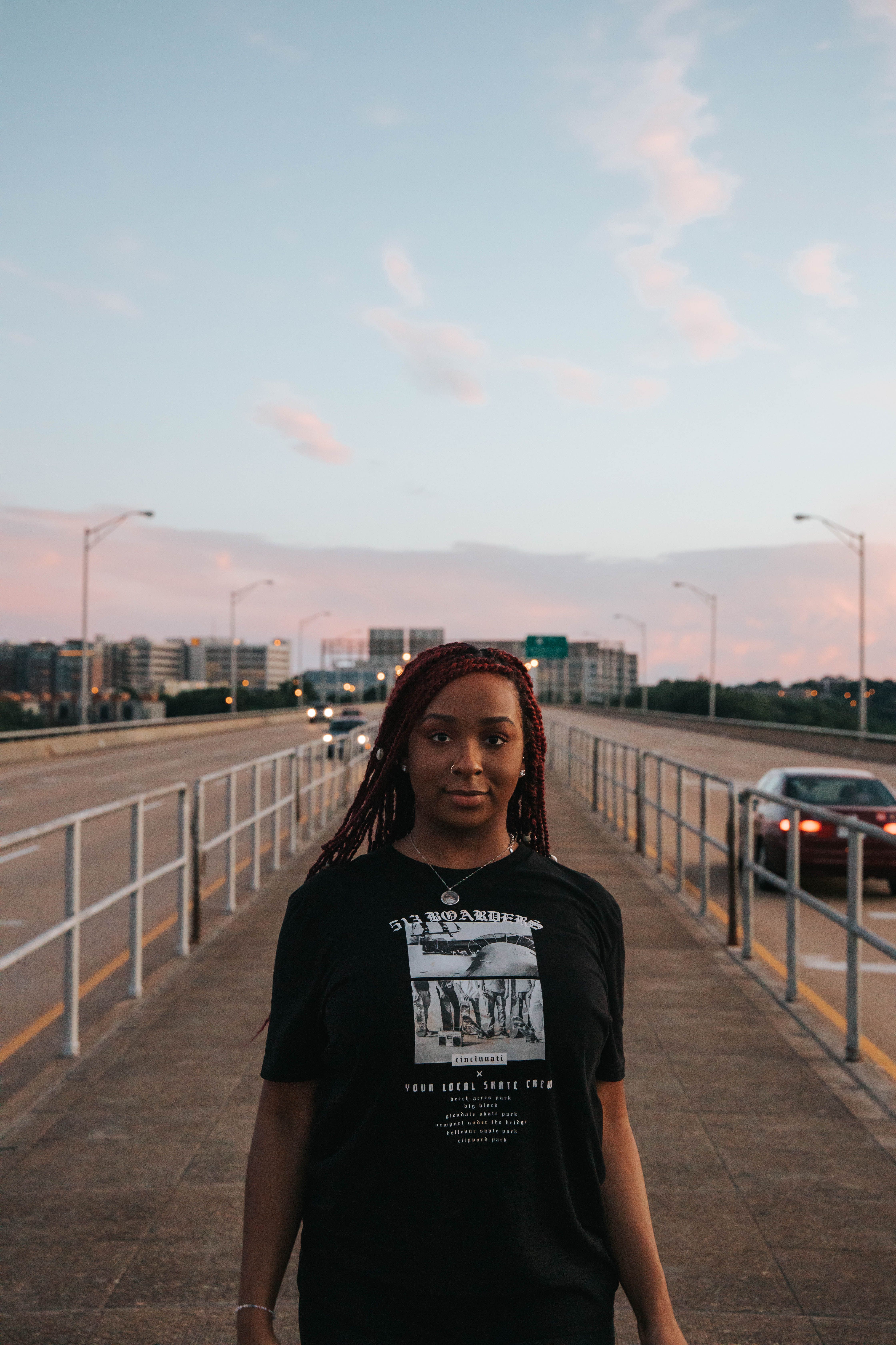 Woman in black t-shirt standing on a bridge with cityscape and cars in the background during sunset.