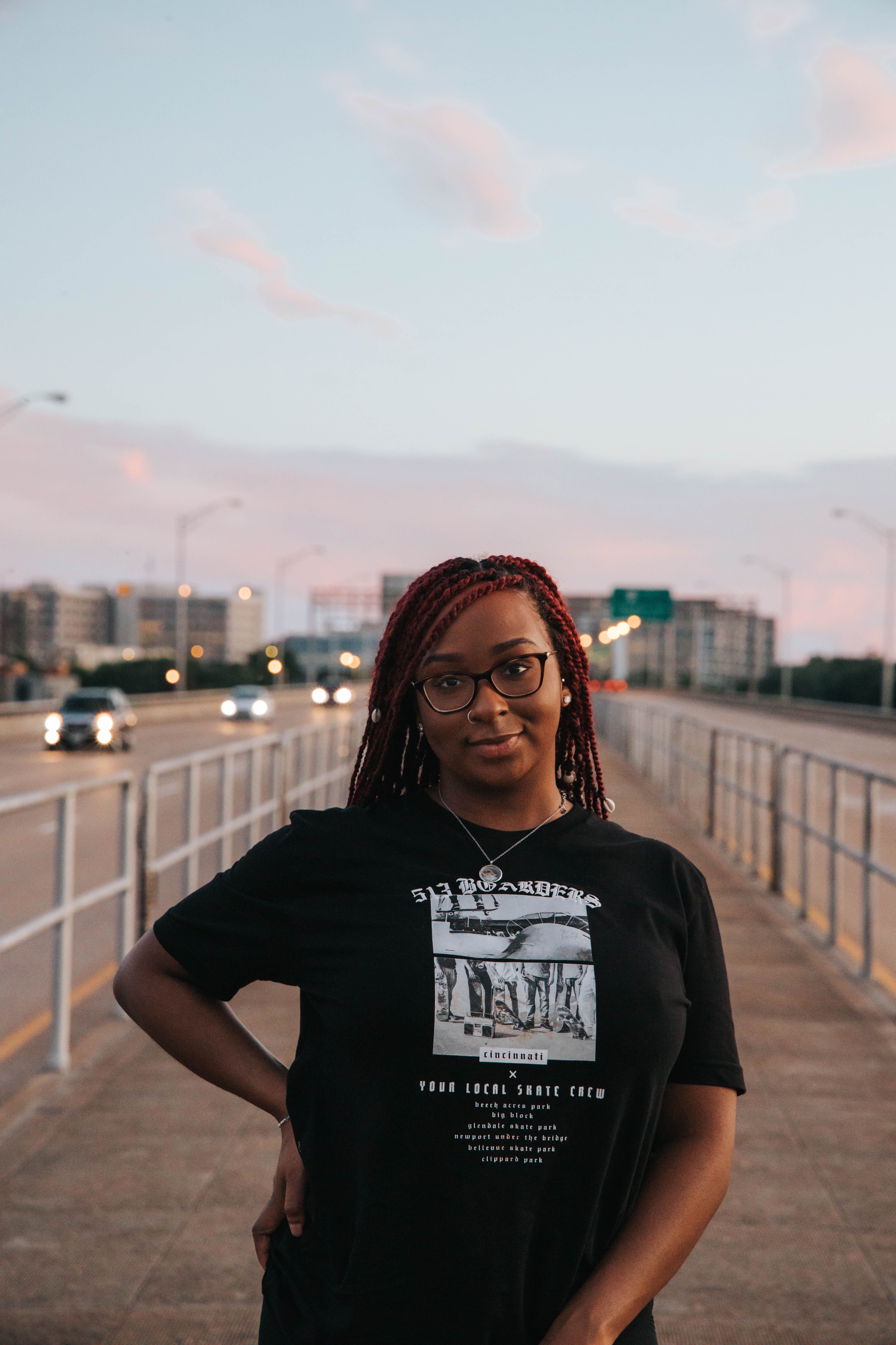 A woman with braided hair and eyeglasses stands confidently on an urban walkway during dusk, with a highway in the background.