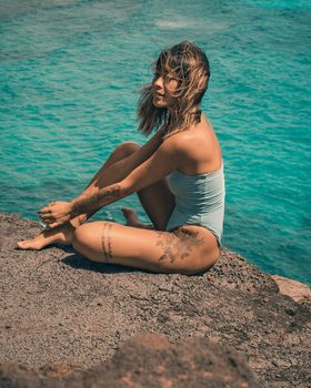 一位穿着泳衣、身上有纹身的女人坐在岩石海岸上，欣赏美景