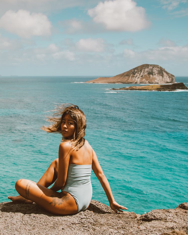 A Woman In One Piece Bikini Seriously Looking At The Camera While Sitting On The Rocky Ground