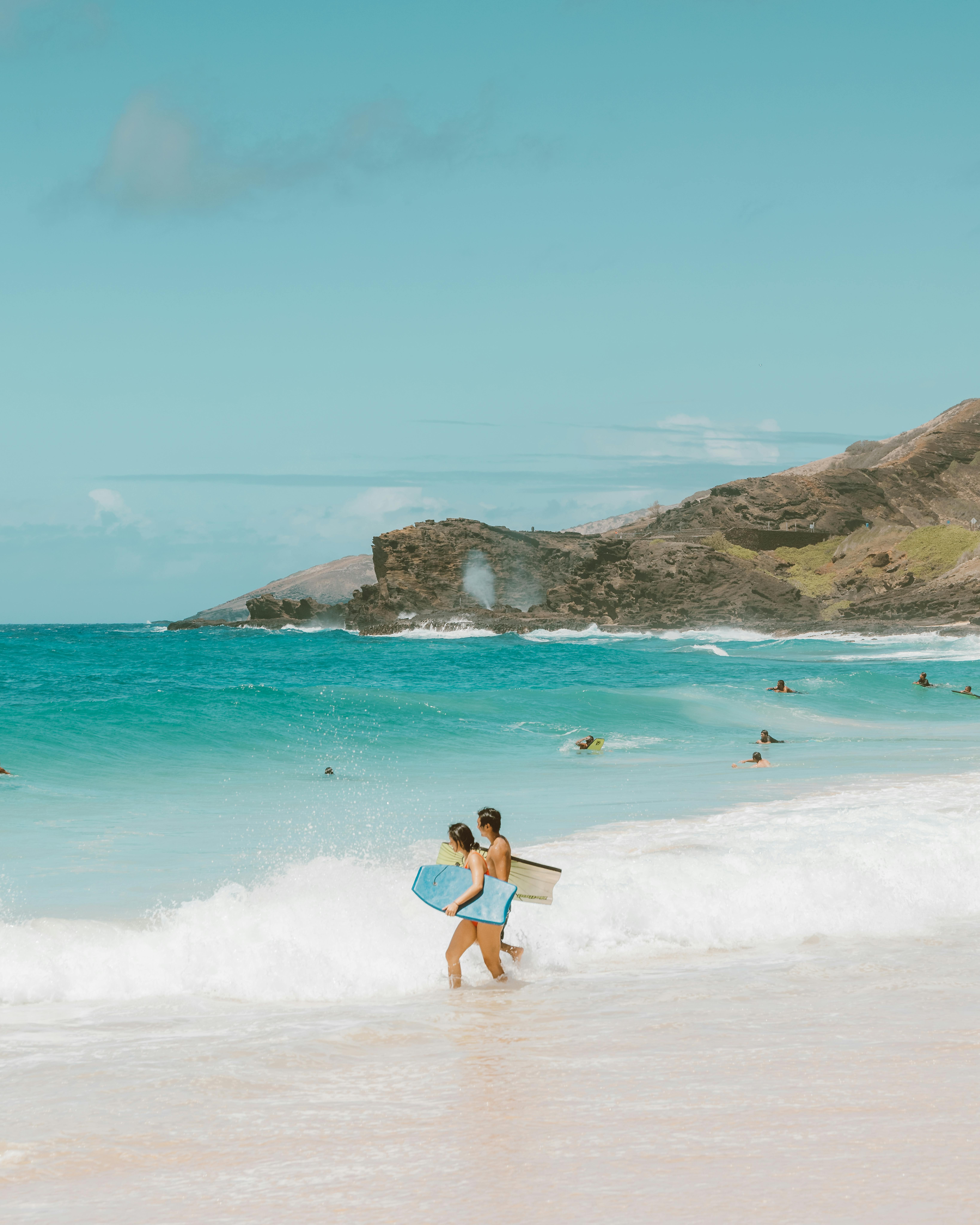 两个冲浪者拿着冲浪板，走在风景秀丽的海滩上，眼前是绿松石般的海浪和郁郁葱葱的山丘