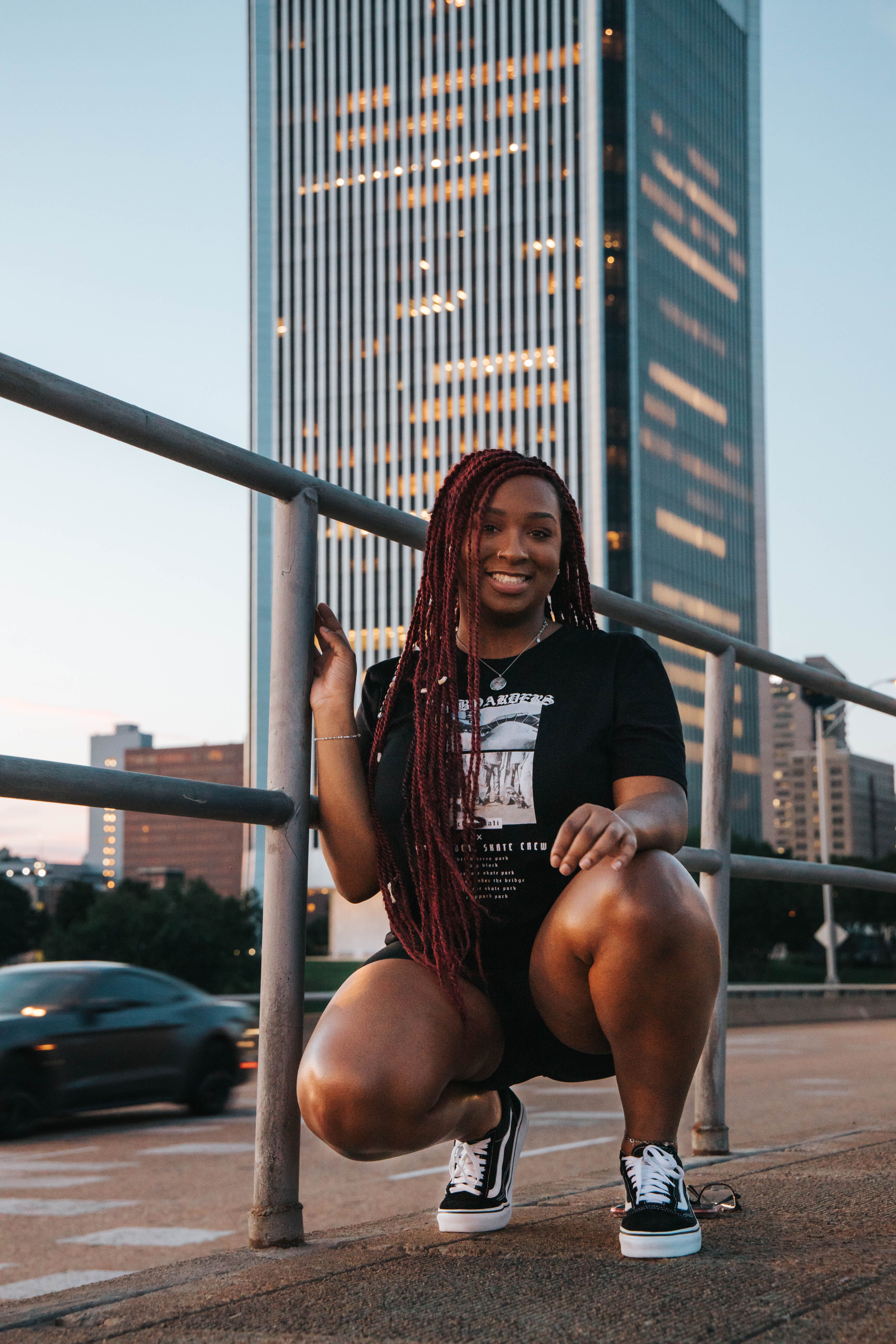 Confident woman kneeling on city street at dusk with tall building background.