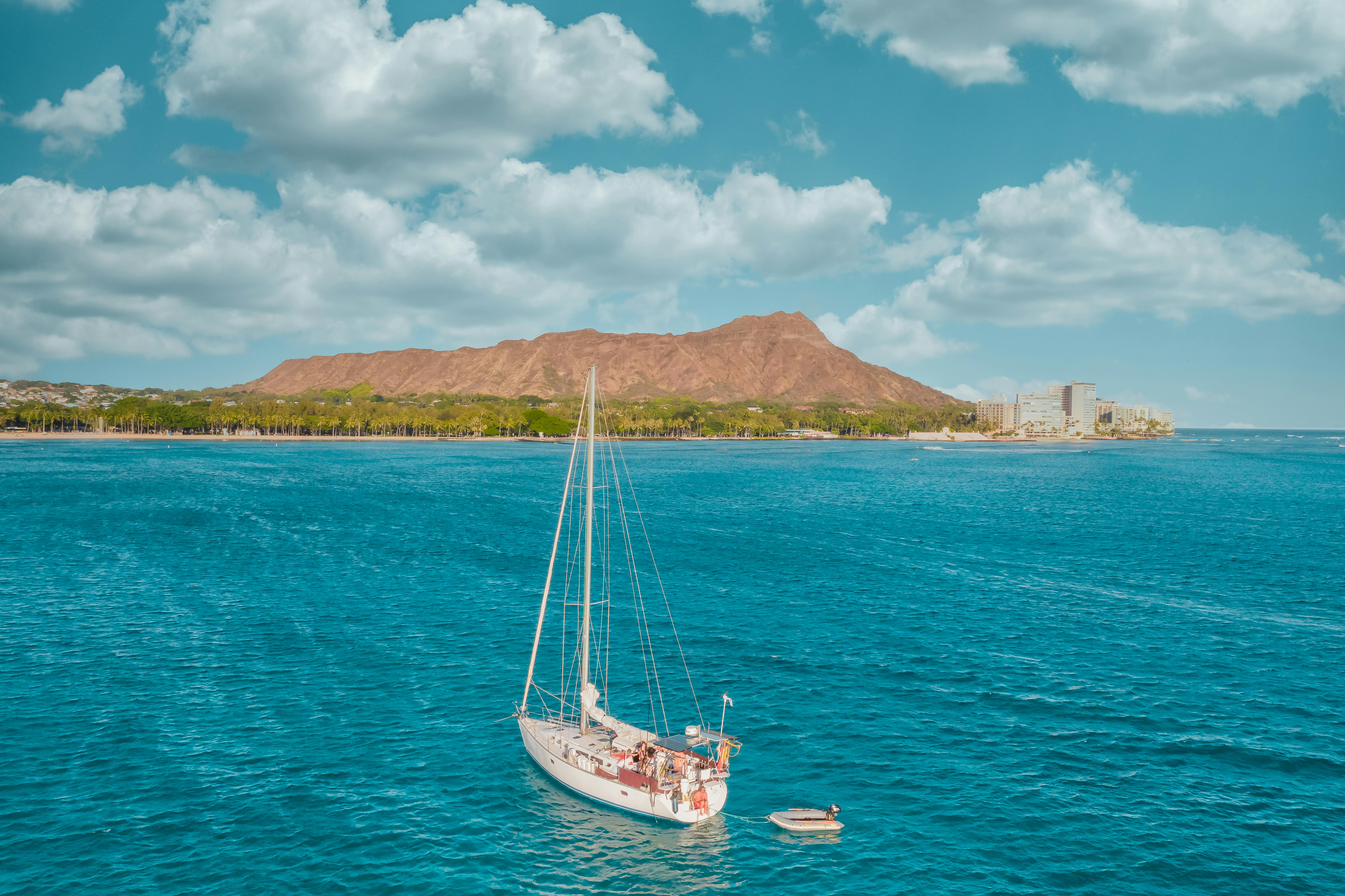 A sailboat cruises in the vivid blue waters near Diamond Head, Hawaii, under clear skies.