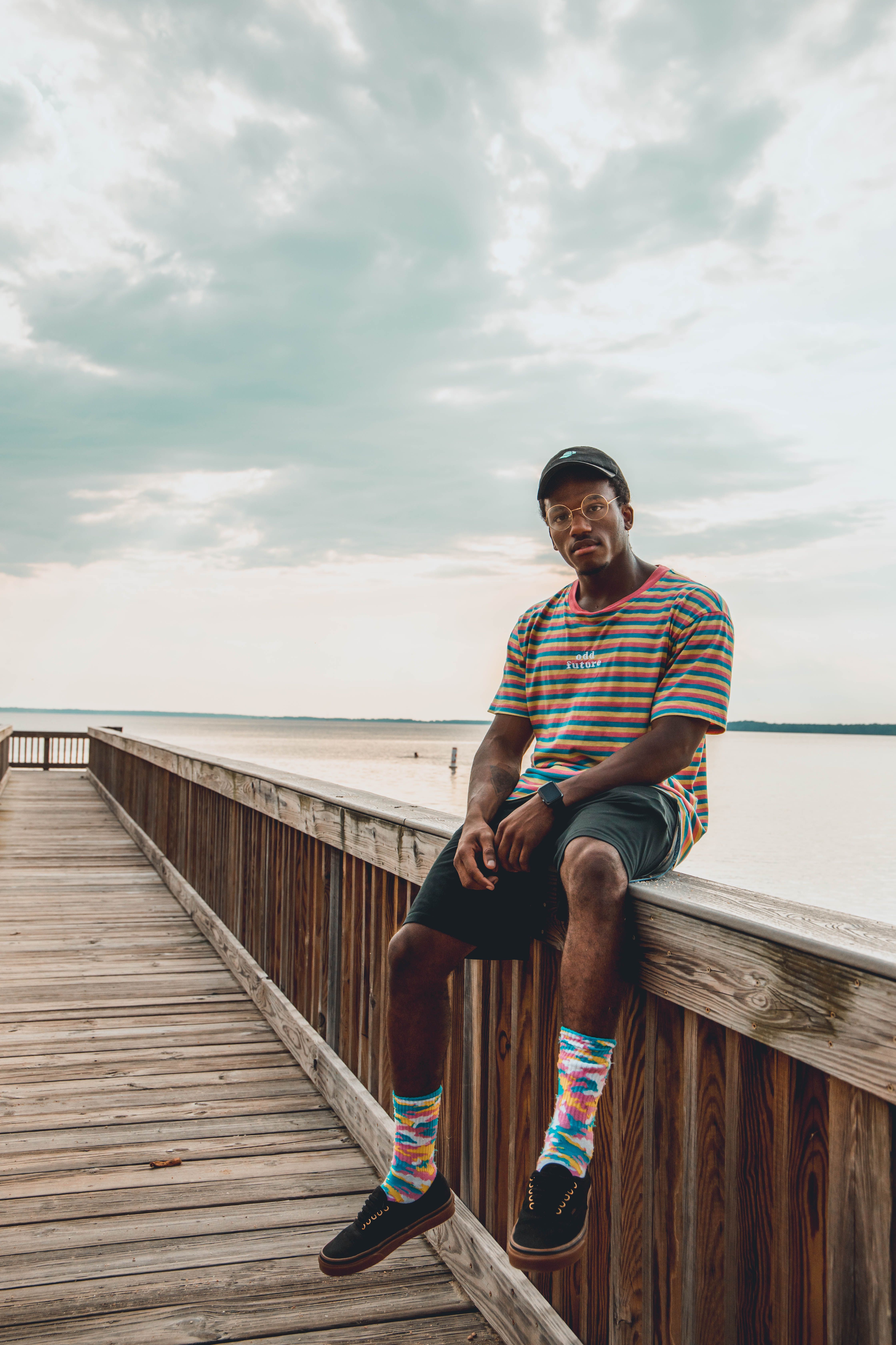 Man wearing colorful socks sits on a wooden dock rail, with a serene lake background under a cloudy sky.