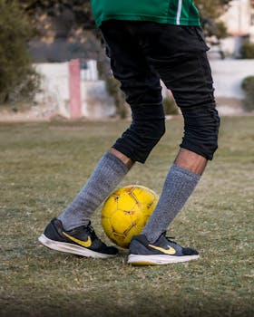A soccer player in action kicking a yellow ball on a grassy field outdoors.