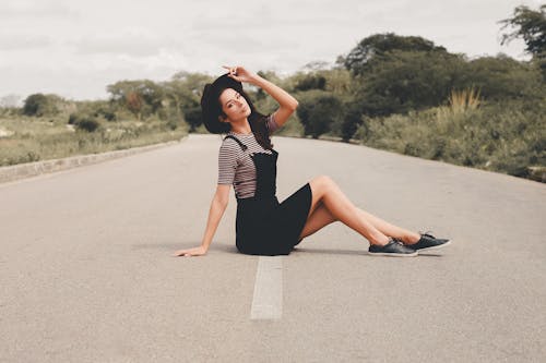 Side view of trendy female sitting on asphalt and looking at camera on summer day in countryside
