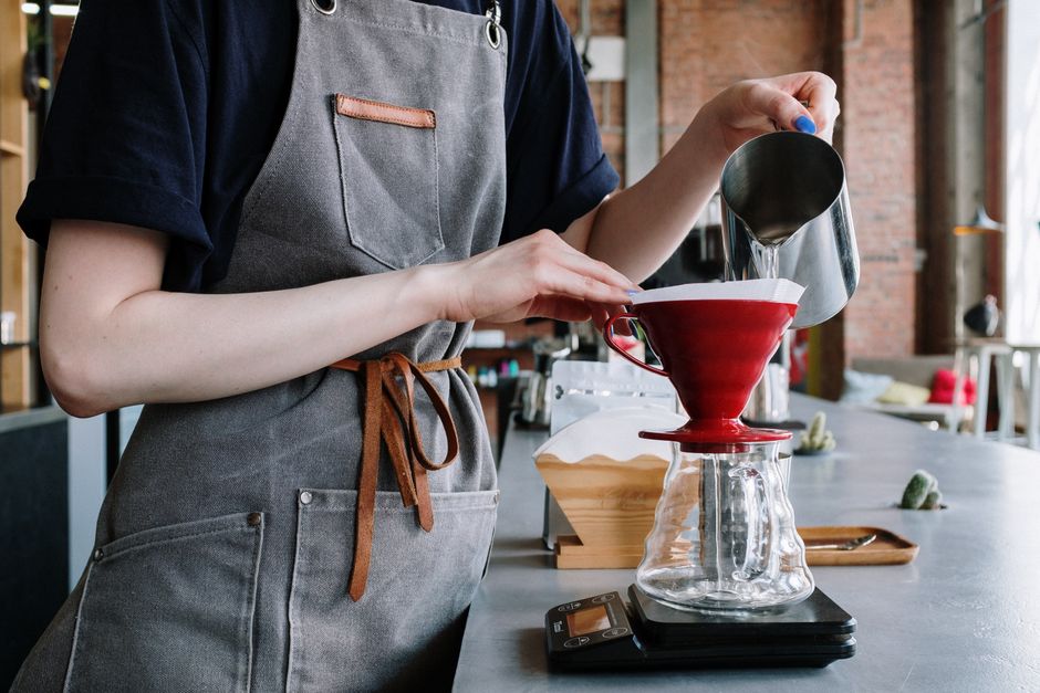 A barista in an apron prepares pour-over coffee using a red funnel and glass carafe.
