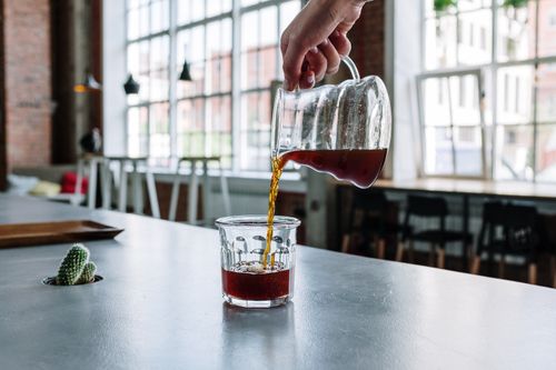 Free A hand pours freshly brewed coffee into a glass in a modern cafe with large windows. Stock Photo