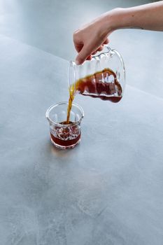 一只手将新鲜咖啡从玻璃罐中倒入放在光滑灰色表面的杯子里