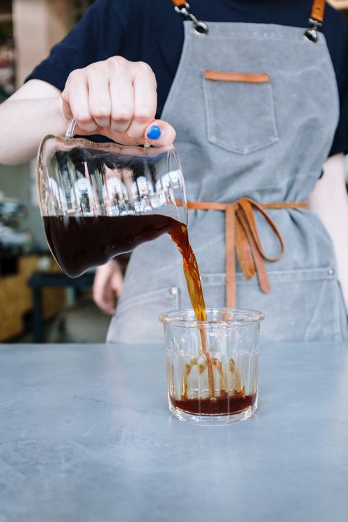 Free Barista in a grey apron pouring freshly brewed coffee into a glass cup indoors. Stock Photo