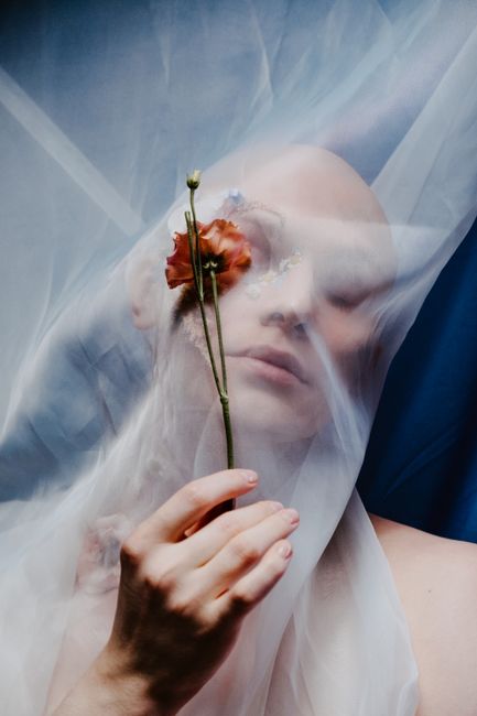 Bald person in an ethereal portrait with translucent veil and floral accents, capturing beauty and sophistication.