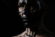 在黑暗背景下，一个脸上涂着大胆黑色油彩的男人特写，传达出独特的艺术表达