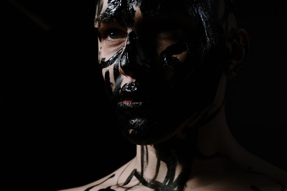 Close-up of a man in bold black face paint against a dark background, conveying a unique and artistic expression.