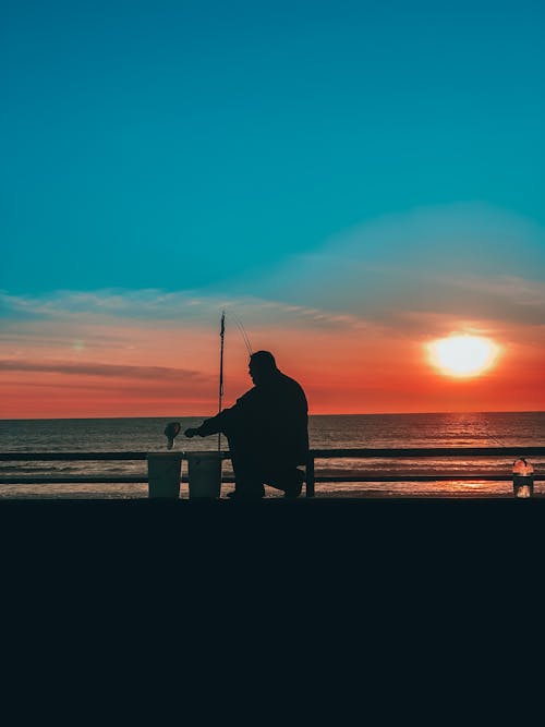 Free Scenic view of male silhouette sitting on peaceful seacoast with rod and fishing against amazing sunset sky in twilight Stock Photo