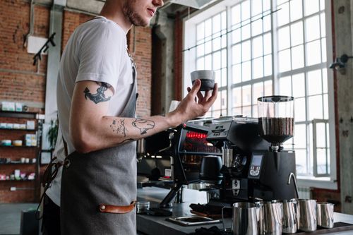 Free Barista in modern cafe holding coffee cup beside espresso machine, showcasing coffee culture. Stock Photo