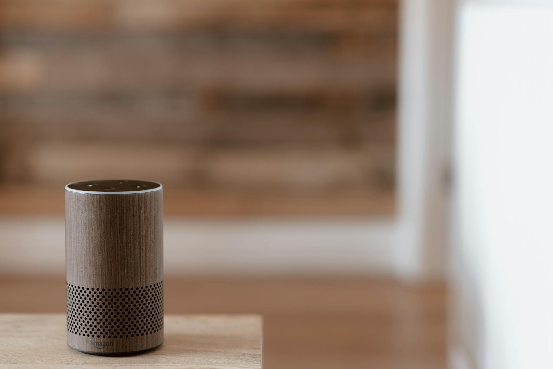 A stylish smart speaker sits on a wooden surface in a cozy, warm indoor setting.