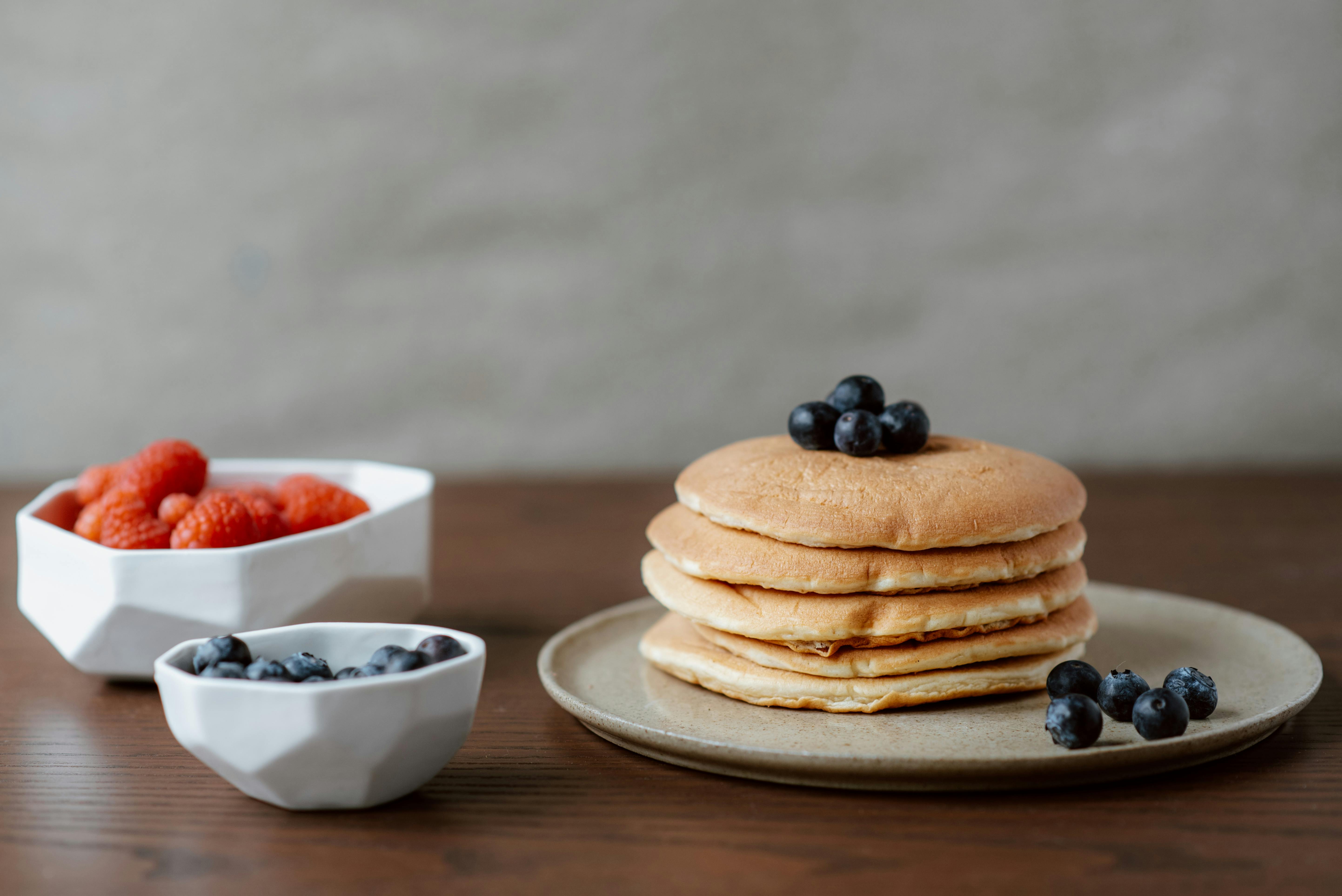 Delicious stack of pancakes served with blueberries and raspberries in white bowls on a wooden table.