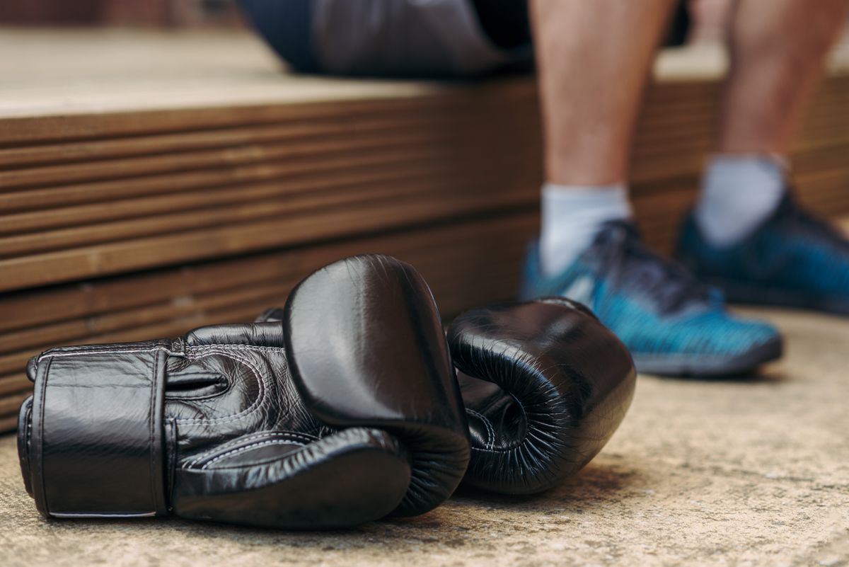 Schwarze Boxhandschuhe liegen auf dem Hallenboden, dahinter die Füße eines Sportlers