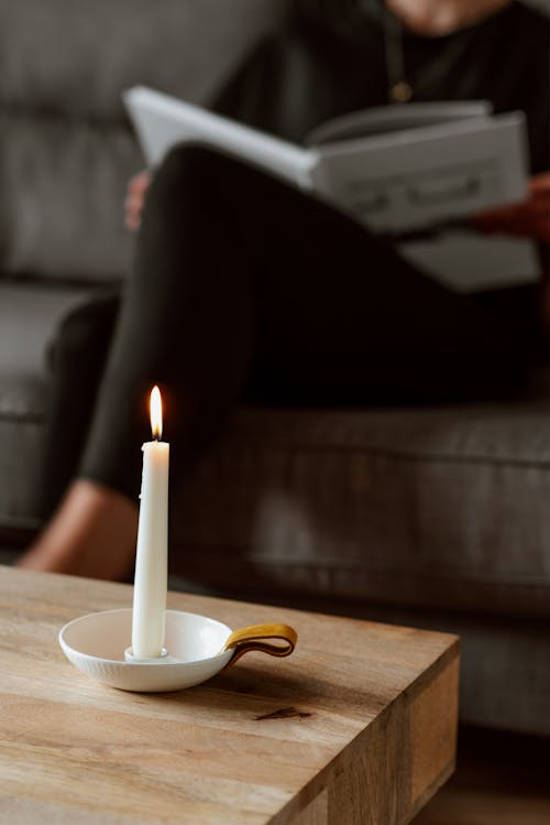Free Crop anonymous female reading textbook with crossed legs on couch behind glowing candle on table at home Stock Photo