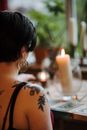 Back view of crop anonymous female soothsayer with tattoos in earring predicting fate with tarot cards near burning candles