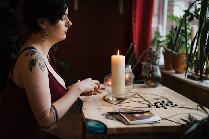 Side view of crop tattooed female soothsayer at table with glowing candle and tarot cards during divination process at home