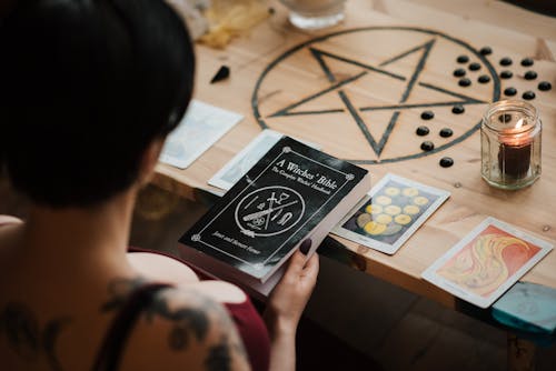 From above back view of crop unrecognizable female soothsayer with magic book at table with tarot cards at home