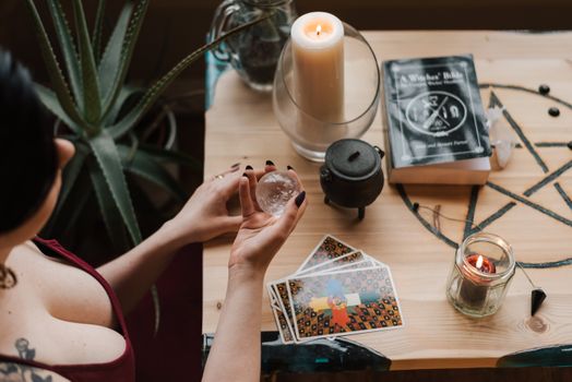 From above of crop unrecognizable female soothsayer with magic ball near tarot cards and candles during divination process in house