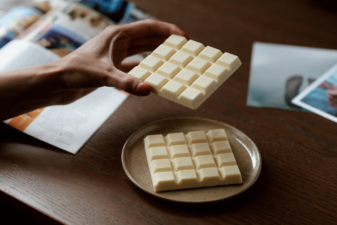 Crop faceless female holding piece of white chocolate while reading magazine and resting at home